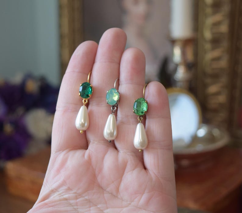 May include: Three pairs of elegant drop earrings. Each earring features a gold-colored hook, a green gemstone, and a pear-shaped faux pearl. The earrings are displayed against a soft, blurred background, highlighting their delicate design.