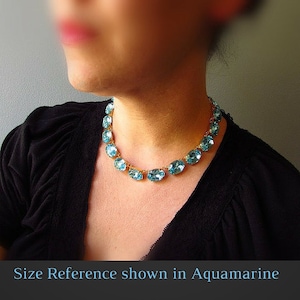 May include: A gold chain necklace with oval-shaped light blue gemstones. The necklace is shown on a model wearing a black shirt. "Size Reference shown in Aquamarine" is written at the bottom of the image.