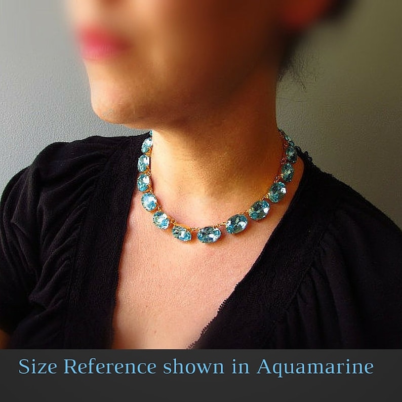 May include: A gold chain necklace with oval-shaped light blue gemstones. The necklace is shown on a model wearing a black shirt. "Size Reference shown in Aquamarine" is written at the bottom of the image.