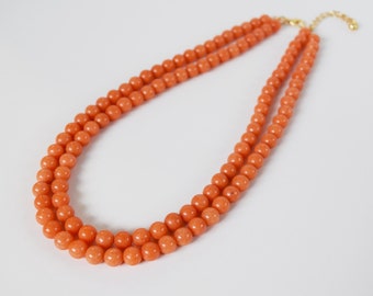 May include: A double-strand necklace featuring orange-colored, round beads. The necklace has a gold-colored clasp and extender chain. The beads are evenly sized and closely spaced, creating a smooth, continuous line of color.