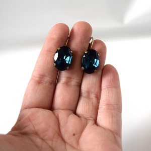 Puede incluir: Un par de pendientes ovalados azul oscuro con ganchos dorados. Los pendientes se sostienen en una mano sobre un fondo blanco. Los pendientes tienen un aspecto facetado, reflejando la luz.