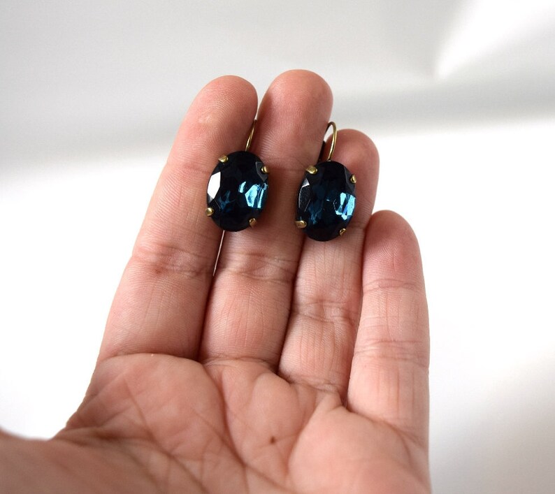 Puede incluir: Un par de pendientes ovalados azul oscuro con ganchos dorados. Los pendientes se sostienen en una mano sobre un fondo blanco. Los pendientes tienen un aspecto facetado, reflejando la luz.
