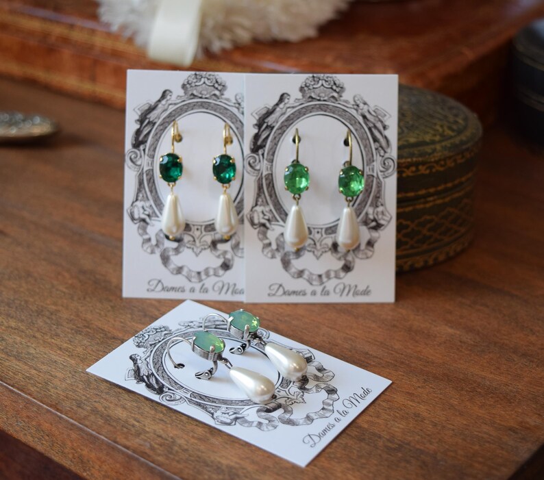 May include: Elegant green and white drop earrings with a pearlescent teardrop shape. The earrings have a gold-colored hook and are displayed on a card with the text "Dames a la Mode".