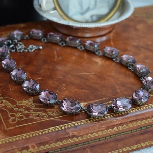 May include: A purple crystal necklace with oval-shaped stones set in silver metal. The necklace is displayed on a brown leather-bound book.