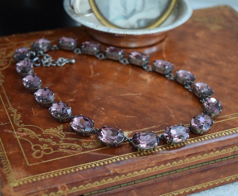 May include: A purple crystal necklace with oval-shaped stones set in silver metal. The necklace is displayed on a brown leather-bound book.