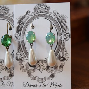 May include: Close-up of three pairs of dangle earrings. Each earring features a colored oval gemstone, either green or light green, and a pear-shaped white faux pearl. The earrings are displayed on cards with decorative illustrations.