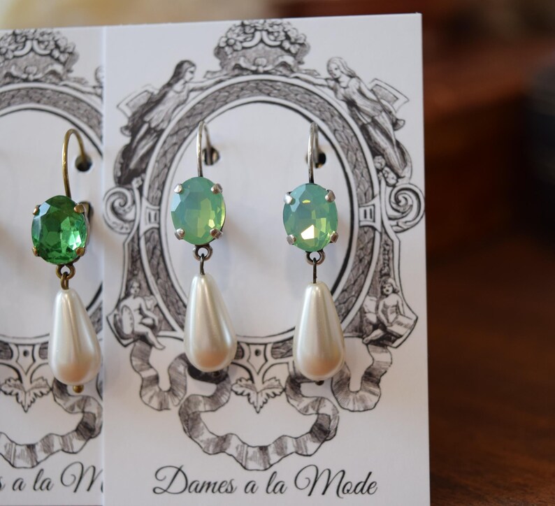 May include: Close-up of three pairs of dangle earrings. Each earring features a colored oval gemstone, either green or light green, and a pear-shaped white faux pearl. The earrings are displayed on cards with decorative illustrations.