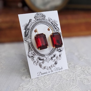 May include: A pair of gold dangle earrings with red rectangular gemstones. The earrings are displayed on a white card with the text "Dames a la Mode".