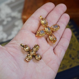 May include: Two gold tone cross shaped pendants with clear amber colored rhinestones. The crosses are on a hand.