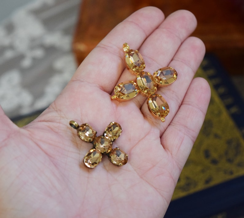 May include: Two gold tone cross shaped pendants with clear amber colored rhinestones. The crosses are on a hand.