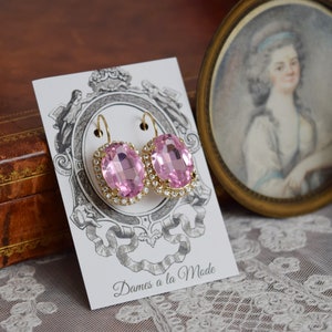 Puede incluir: Un par de pendientes de oro con piedras ovaladas de color rosa engastadas en un halo de brillantes transparentes. Los pendientes se muestran en una tarjeta blanca con el texto "Dames a la Mode".