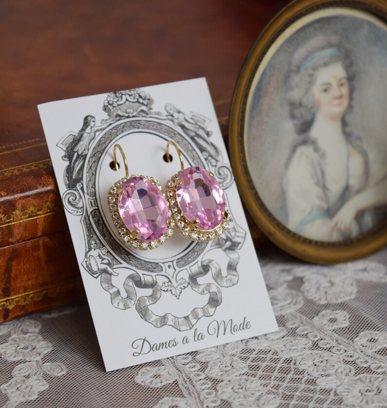 Puede incluir: Un par de pendientes de oro con piedras ovaladas de color rosa engastadas en un halo de brillantes transparentes. Los pendientes se muestran en una tarjeta blanca con el texto "Dames a la Mode".