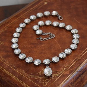 May include: A silver necklace with a teardrop pendant made of clear faceted crystals. The necklace is on a brown leather surface.
