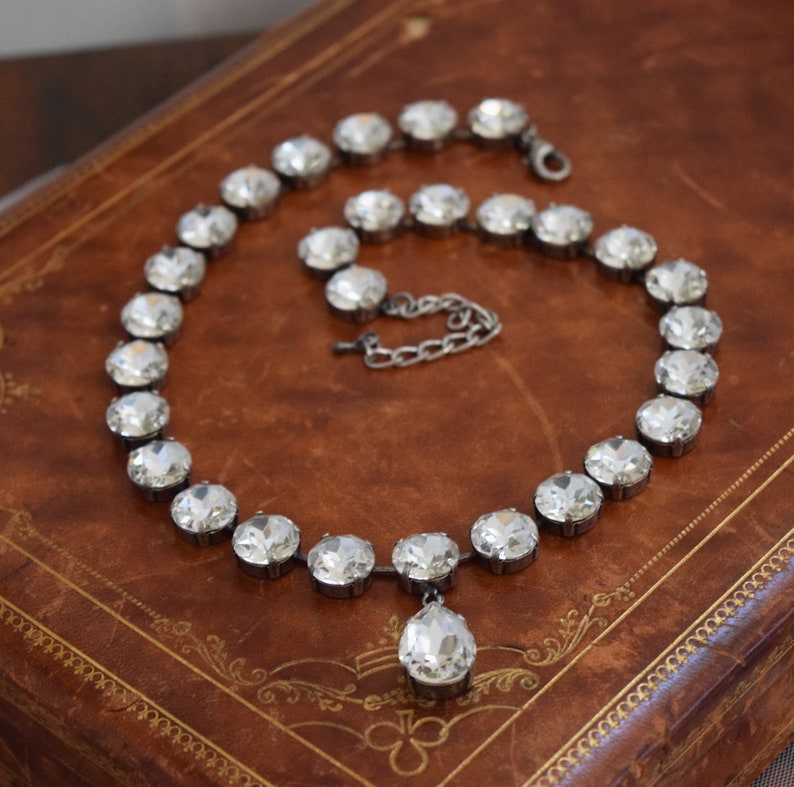 May include: A silver necklace with a teardrop pendant made of clear faceted crystals. The necklace is on a brown leather surface.