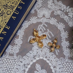 May include: Two gold-toned, crystal-like flower charms with a delicate design. The charms are on a white lace background.