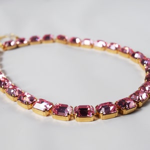 May include: A pink rhinestone choker necklace with gold-toned metal settings. The necklace is made of rectangular-shaped rhinestones.