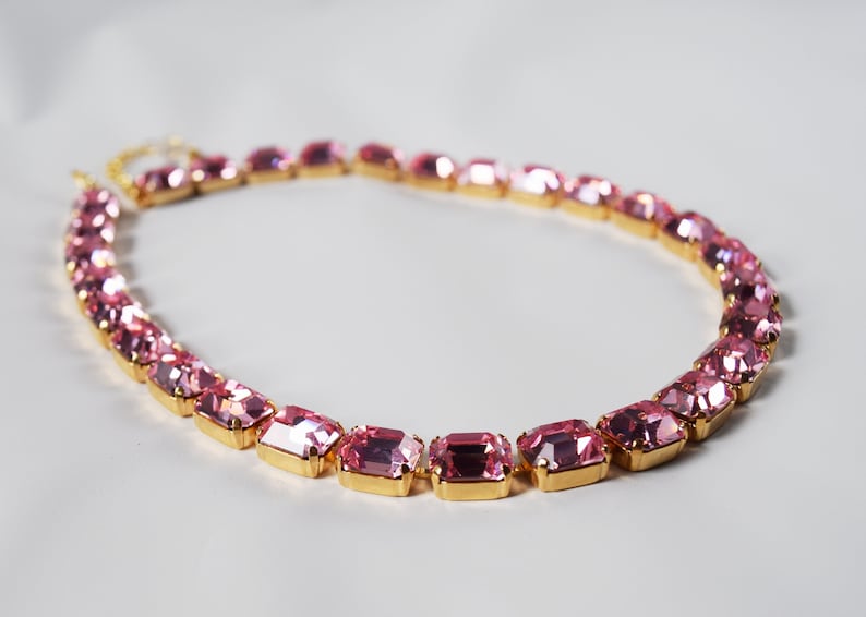 May include: A pink rhinestone choker necklace with gold-toned metal settings. The necklace is made of rectangular-shaped rhinestones.