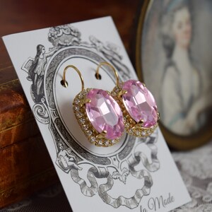 Puede incluir: Un par de pendientes ovalados de color rosa con engastes dorados y un acabado cristalino. Los pendientes est&aacute;n sobre una tarjeta blanca con un dise&ntilde;o decorativo.