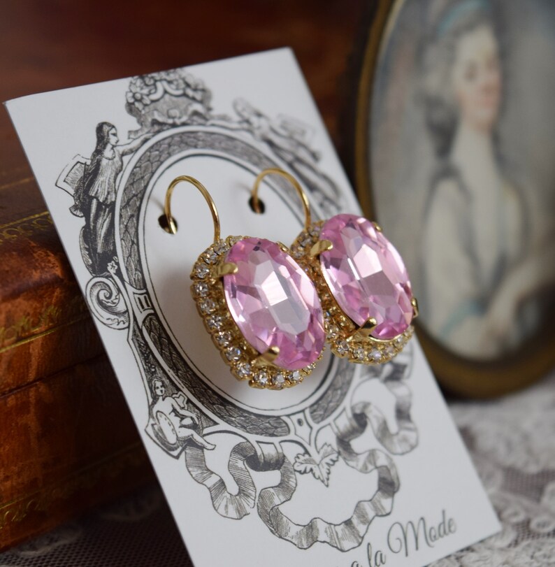 Puede incluir: Un par de pendientes ovalados de color rosa con engastes dorados y un acabado cristalino. Los pendientes est&aacute;n sobre una tarjeta blanca con un dise&ntilde;o decorativo.