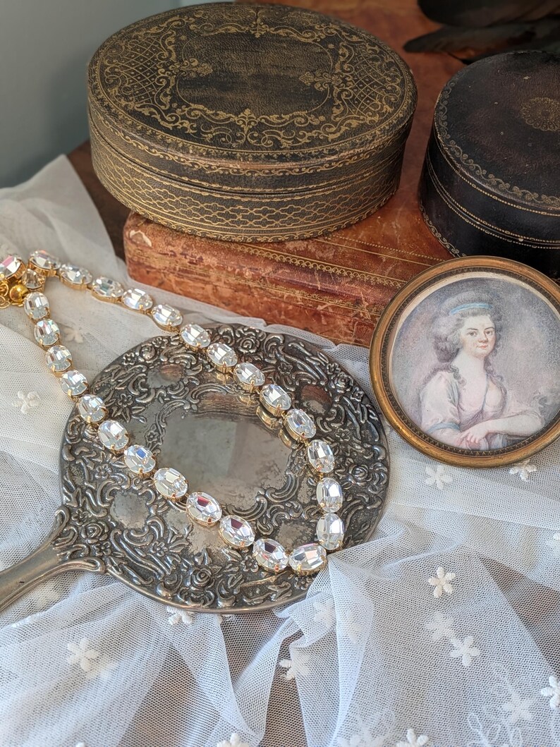 Puede incluir: Un collar de tono dorado que presenta una serie de cristales transparentes de forma ovalada y facetados, unidos entre s&iacute;. El collar se muestra sobre un espejo de mano redondo de color plateado con intrincados relieves florales.