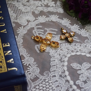 May include: Two gold-toned, cross-shaped, rhinestone embellishments with amber-colored stones. The embellishments are on a white lace fabric background.