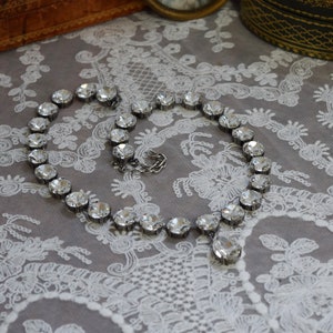 May include: A silver necklace with a teardrop pendant and clear crystal beads. The necklace is lying on a white lace fabric.