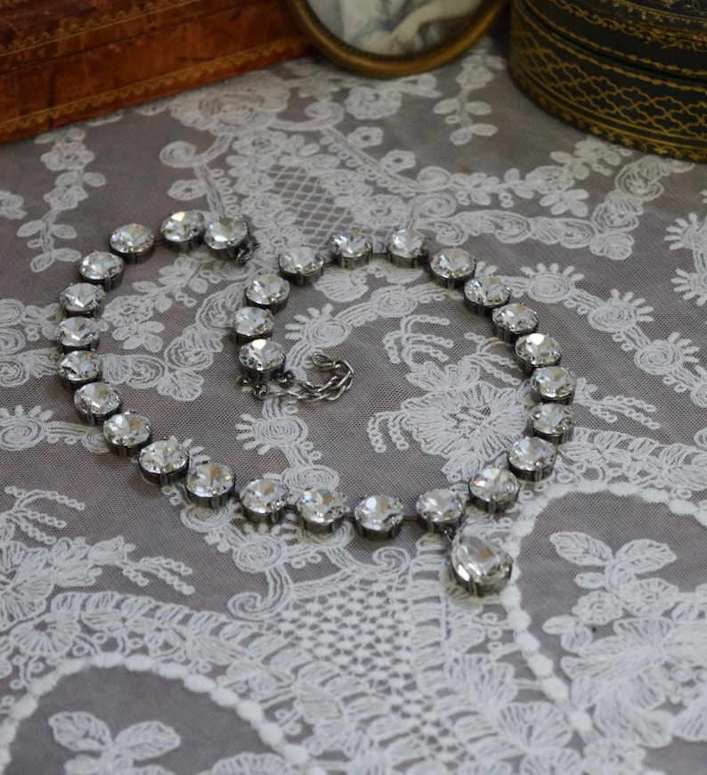 May include: A silver necklace with a teardrop pendant and clear crystal beads. The necklace is lying on a white lace fabric.