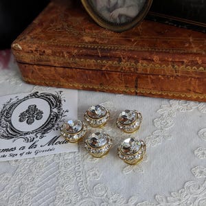 May include: Five gold-toned, round, crystal-encrusted buttons with clear rhinestones. The buttons are arranged in a semi-circle on a white lace surface.