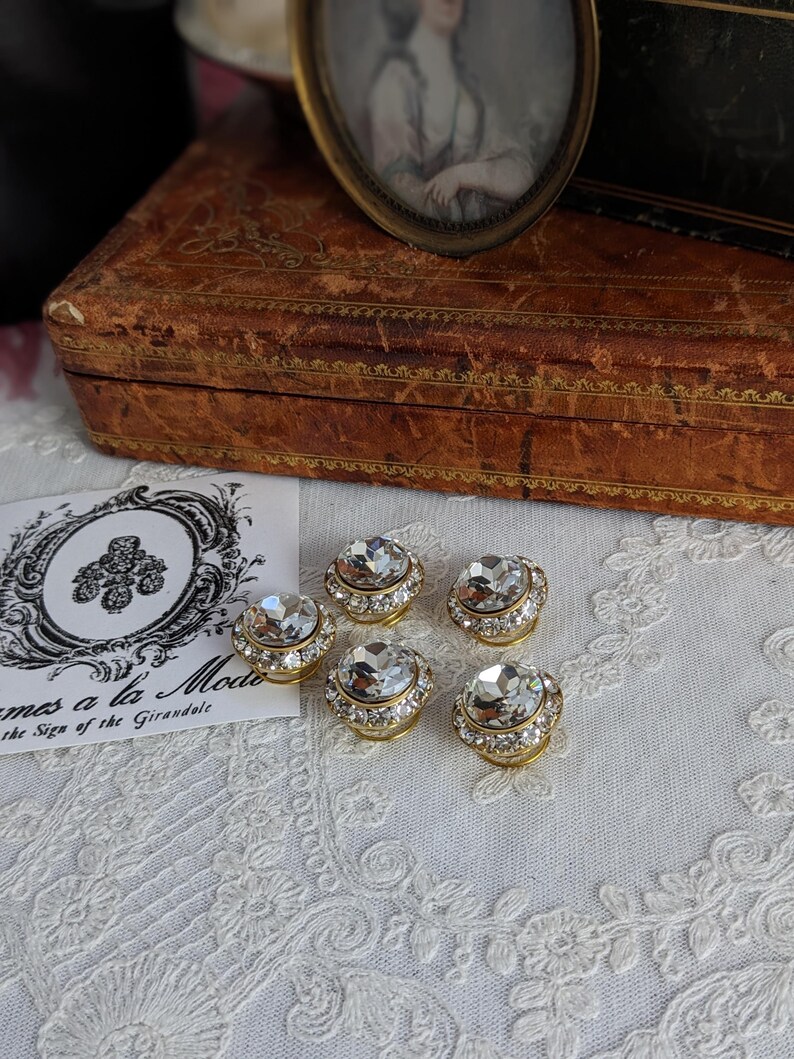 May include: Five gold-toned, round, crystal-encrusted buttons with clear rhinestones. The buttons are arranged in a semi-circle on a white lace surface.
