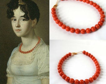 May include: A close-up of two red coral necklaces. The necklaces are made of round beads and are strung on a gold chain. The necklaces are displayed on a white background.