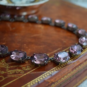 May include: A silver chain necklace with large oval-shaped amethyst-coloured rhinestones. The necklace is lying on a brown leather surface.