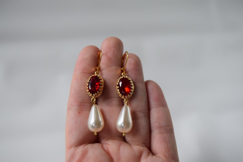 May include: Gold earrings with a red gemstone and a white pearl drop.