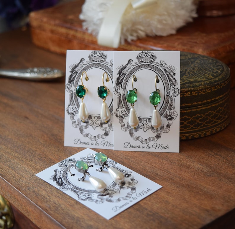 May include: Elegant drop earrings with green gemstone accents and pear-shaped white faux pearls. The earrings are displayed on cards with the text "Dames a la Mode" and a decorative illustration. The jewelry is set against a wooden surface.