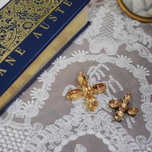 May include: A close-up of two gold-toned, jeweled cross-shaped charms. The charms are on a white lace background. The larger charm has a more intricate design with multiple facets. The smaller charm has a simpler design with fewer facets.