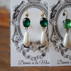 May include: Gold-toned earrings with emerald green oval-cut gems and pear-shaped faux pearls. The earrings hang from gold hooks. The earrings are displayed on a white card with a black and white illustration and the text "Dames a la Moa".