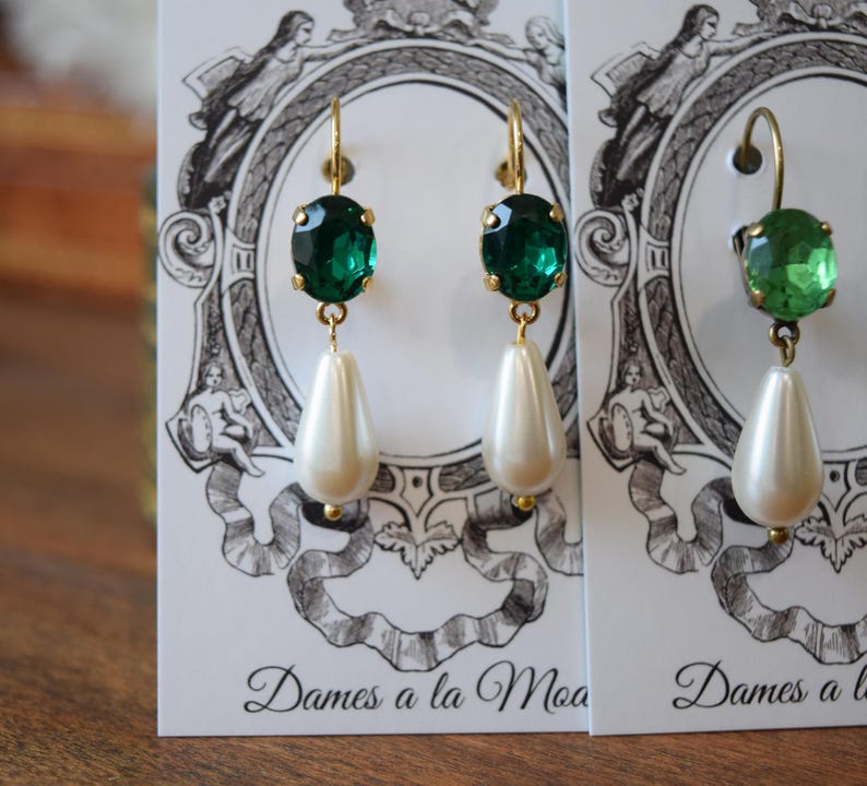 May include: Gold-toned earrings with emerald green oval-cut gems and pear-shaped faux pearls. The earrings hang from gold hooks. The earrings are displayed on a white card with a black and white illustration and the text "Dames a la Moa".