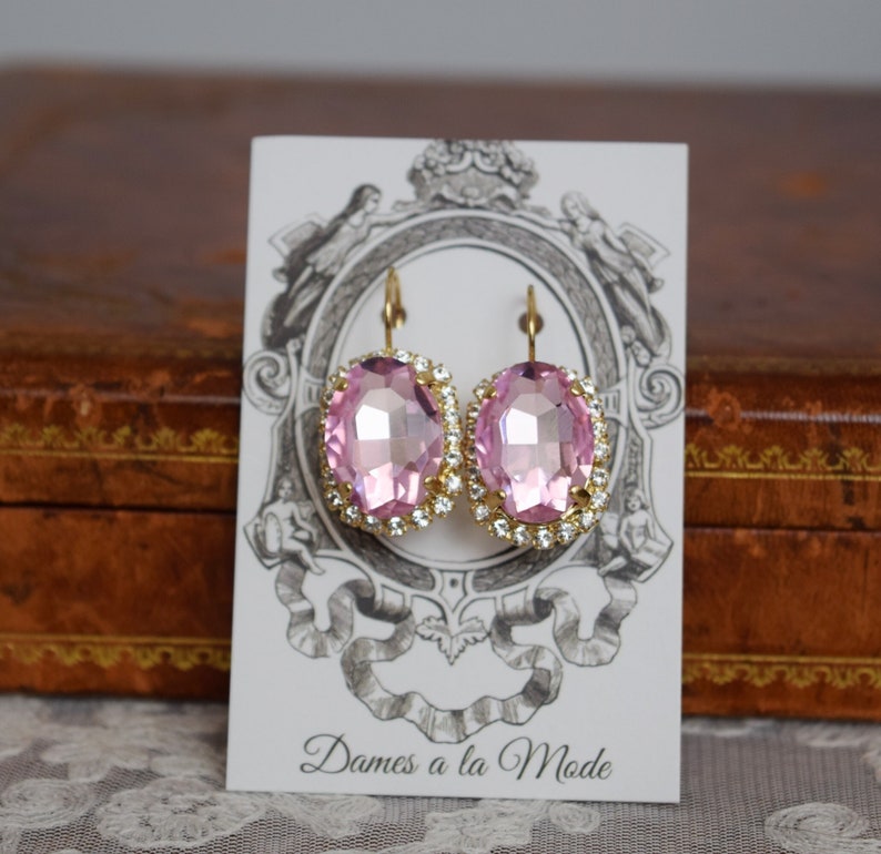 Puede incluir: Un par de pendientes ovalados de color rosa con engastes dorados y un borde de strass transparente. Los pendientes se muestran en una tarjeta blanca con el texto "Dames a la Mode".