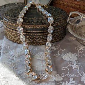Puede incluir: Un collar de tono dorado que presenta una hilera de grandes cristales transparentes facetados de forma ovalada, engarzados en garras de metal. El collar incluye un cierre de metal y un peque&ntilde;o dije circular unido al extremo de la cadena.