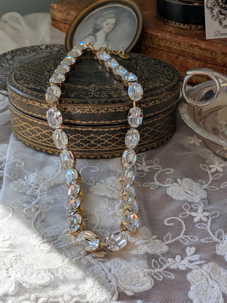 Puede incluir: Un collar de tono dorado que presenta una hilera de grandes cristales transparentes facetados de forma ovalada, engarzados en garras de metal. El collar incluye un cierre de metal y un peque&ntilde;o dije circular unido al extremo de la cadena.