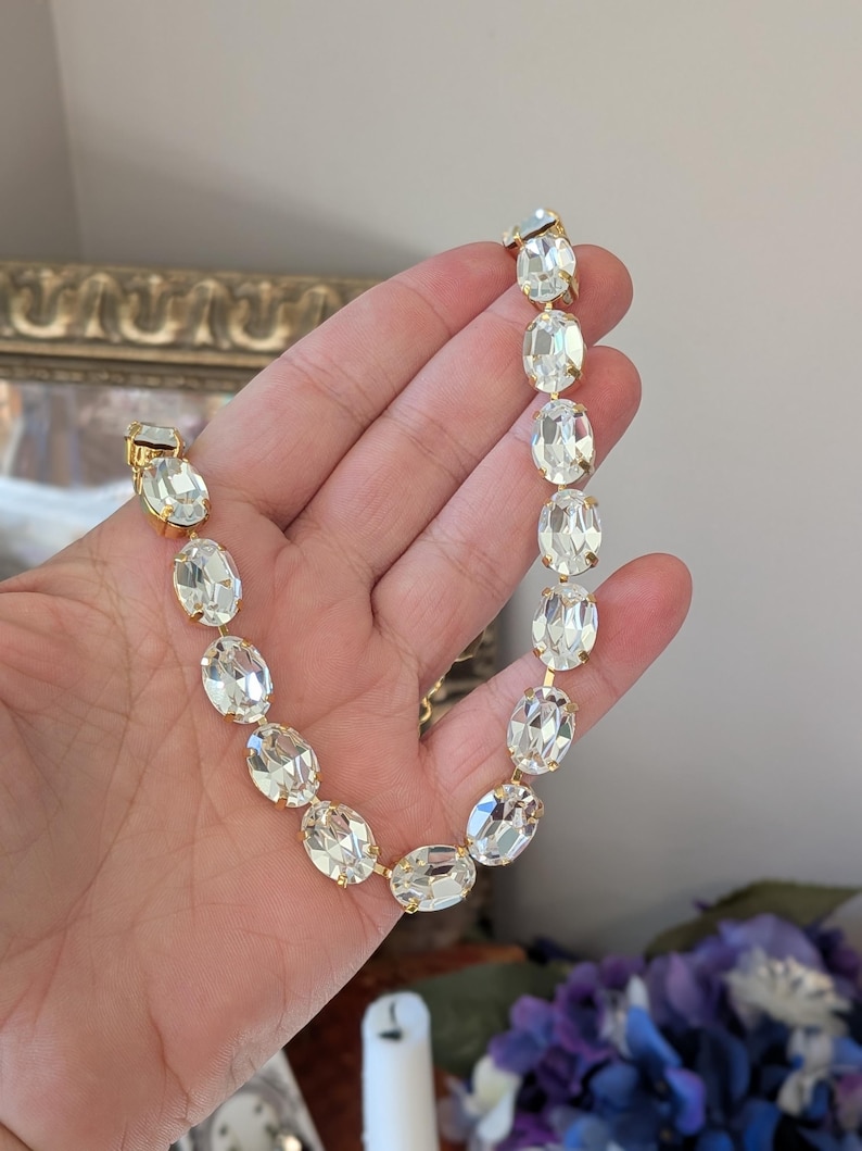 Puede incluir: Un collar de tono dorado que presenta una serie de cristales transparentes de forma ovalada y facetados. Cada cristal est&aacute; montado en un engaste de metal dorado, creando una cadena continua de piedras brillantes que descansa en la palma.
