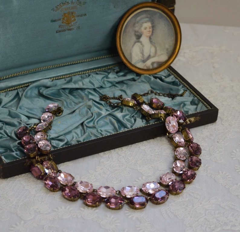 May include: A two-strand necklace made of oval-shaped pink and purple glass stones with antique brass accents. The necklace is displayed in a blue velvet-lined jewelry box.