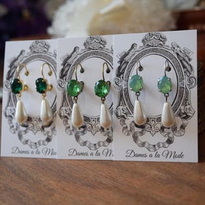May include: Three pairs of elegant earrings displayed on cards. Each pair features a colored gemstone and a dangling pear-shaped faux pearl. The earrings have gold or silver-colored hooks. The cards have a decorative emblem and the text "Dames a la Mode".