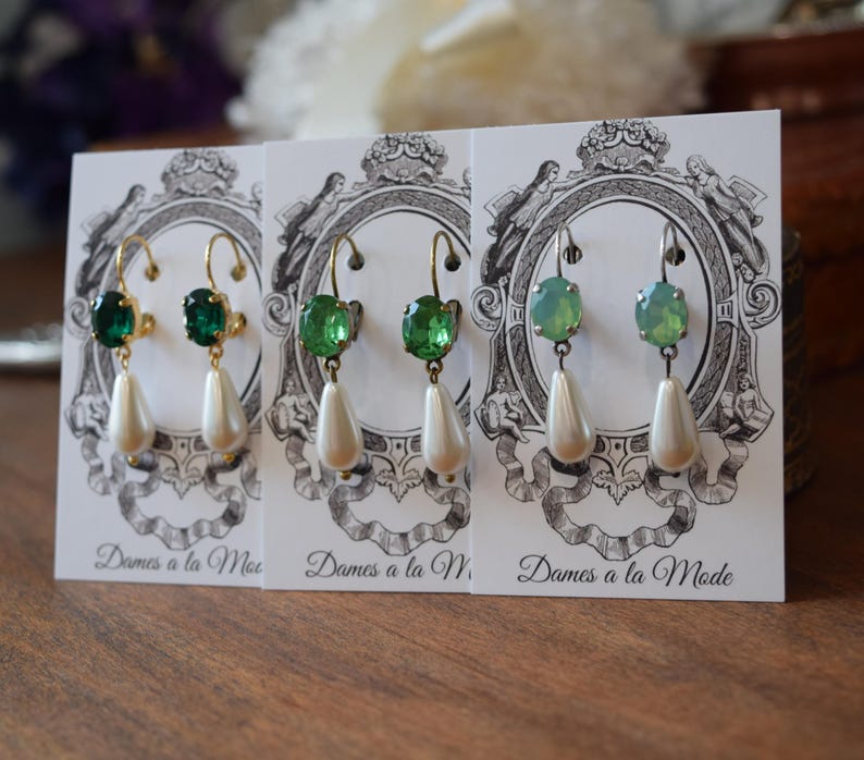 May include: Three pairs of elegant earrings displayed on cards. Each pair features a colored gemstone and a dangling pear-shaped faux pearl. The earrings have gold or silver-colored hooks. The cards have a decorative emblem and the text "Dames a la Mode".
