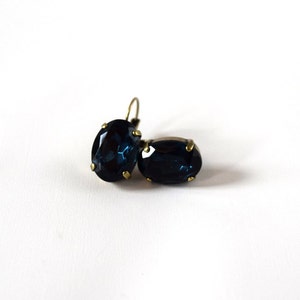 Puede incluir: Un par de pendientes ovalados azul oscuro con engastes y ganchos para las orejas de metal dorado. Los pendientes est&aacute;n sobre un fondo blanco, resaltando su color y forma. Las piedras preciosas tienen una superficie facetada.