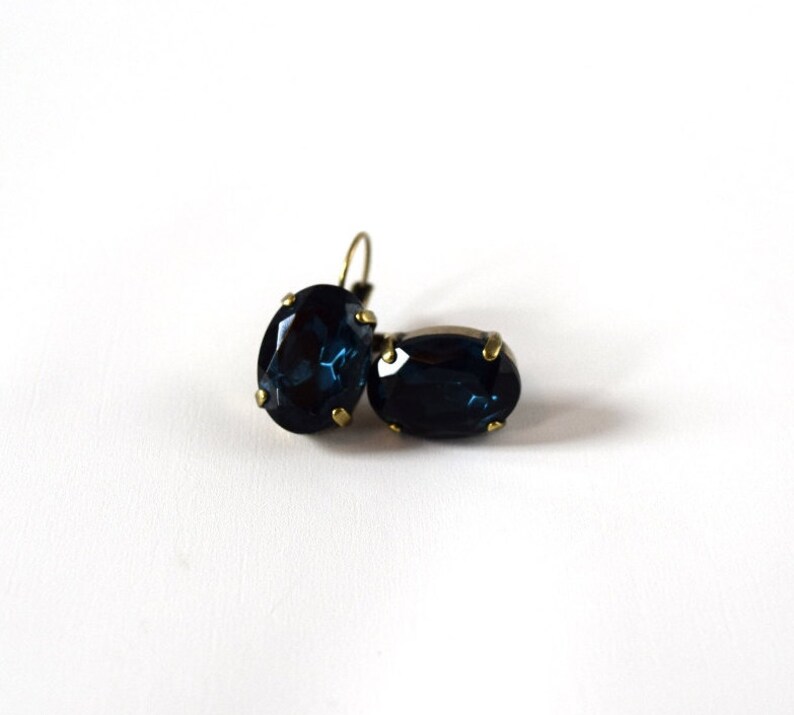 Puede incluir: Un par de pendientes ovalados azul oscuro con engastes y ganchos para las orejas de metal dorado. Los pendientes est&aacute;n sobre un fondo blanco, resaltando su color y forma. Las piedras preciosas tienen una superficie facetada.