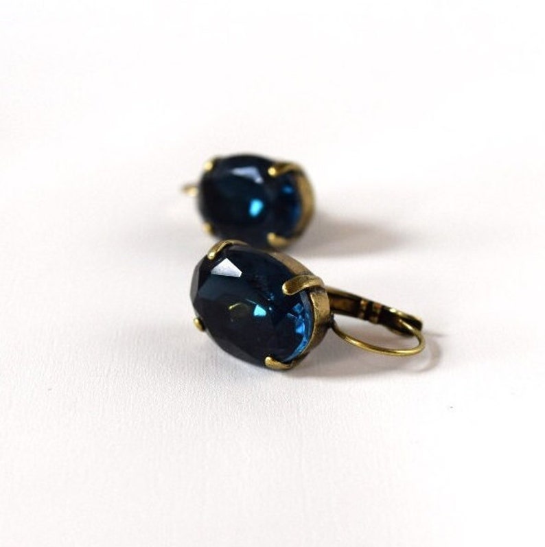 Puede incluir: Un par de pendientes de estilo antiguo con piedras preciosas ovaladas de color azul oscuro. Las piedras preciosas est&aacute;n engastadas en un marco de metal dorado con cierre de palanca. Los pendientes se muestran sobre un fondo blanco.