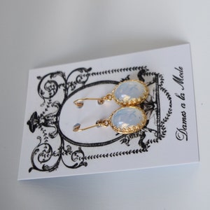 Pode incluir: Um par de brincos de ouro com pedras de opala de forma oval. Os brincos est&atilde;o em um cart&atilde;o branco com o texto "Dames a la Mode".