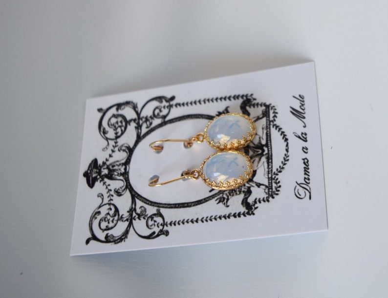 Pode incluir: Um par de brincos de ouro com pedras de opala de forma oval. Os brincos est&atilde;o em um cart&atilde;o branco com o texto "Dames a la Mode".