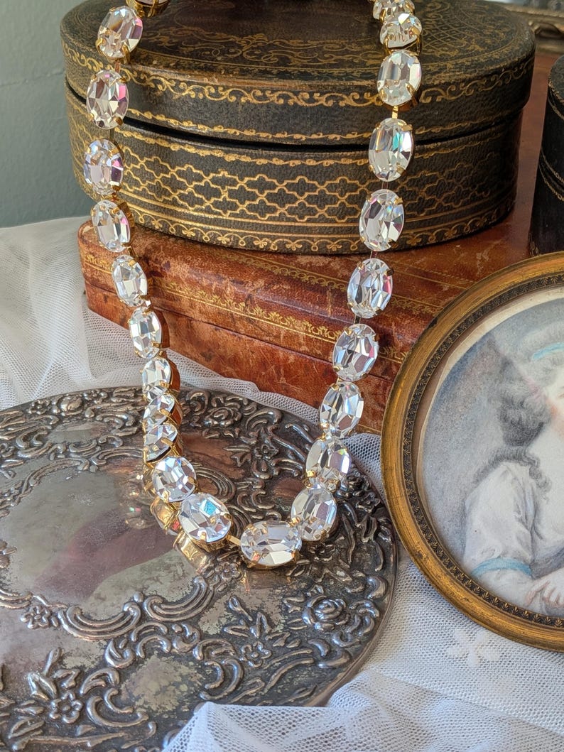 Puede incluir: Un collar que presenta una hilera continua de cristales transparentes de forma ovalada y tallados, montados en engastes de metal dorado. Los cristales est&aacute;n unidos entre s&iacute; formando una cadena flexible que refleja la luz en sus superficies.