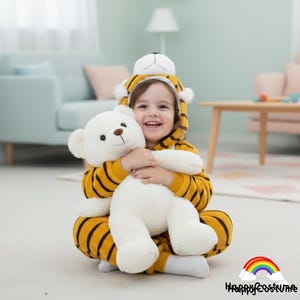 May include: A child wearing a yellow and black tiger-striped jumpsuit with a matching hood, holding a large white teddy bear. The jumpsuit has black stripes and a tiger face on the hood. The child is smiling.
